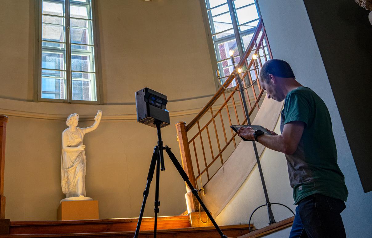 Mann erstellt mit einer Kamera eine 3D-Aufnahme in einem Raum mit einer Holztreppe und großen Fenstern. Stadtmuseum.