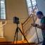 Mann erstellt mit einer Kamera eine 3D-Aufnahme in einem Raum mit einer Holztreppe und großen Fenstern. Stadtmuseum.