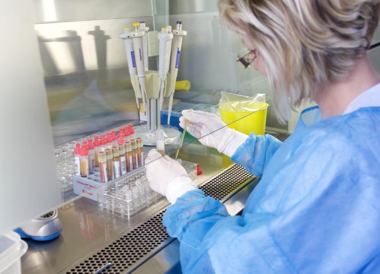 Frau mit Schutzkleidung hält Pipette mit Flüssigkeit in rechter Hand und Röhrchen in linker Hand