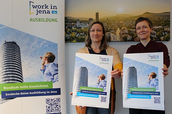 zwei Frauen stehen auf der rechten Seite des Bildes, sehen Betrachter direk an, halten jeweils ein Plakat in den Händen, links neben Ihnen und im Hintergrund sind weitere Plakate zu sehen