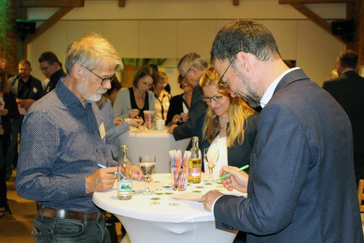 Zwei Männer in Hemd und Anzug stehen an einem Stehtisch und schreiben etwas auf eine Karteikarte.