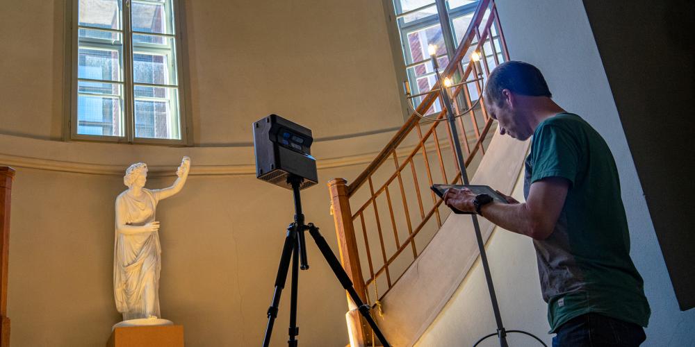 Mann erstellt mit einer Kamera eine 3D-Aufnahme in einem Raum mit einer Holztreppe und großen Fenstern. Stadtmuseum.