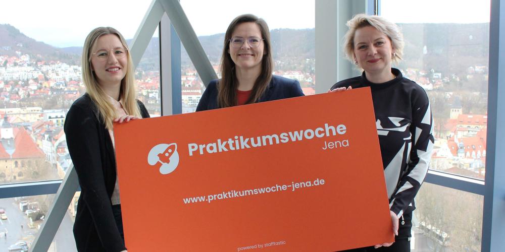 Drei stehen vor großer Fensterfront, stehen nebeneinander und halten oranges Schild mit weißer Schrift