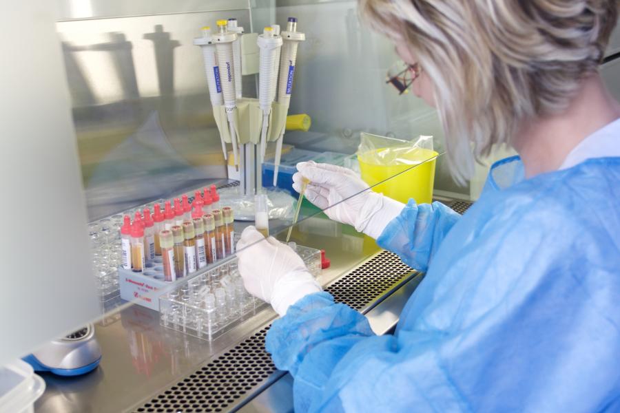Frau mit Schutzkleidung hält Pipette mit Flüssigkeit in rechter Hand und Röhrchen in linker Hand