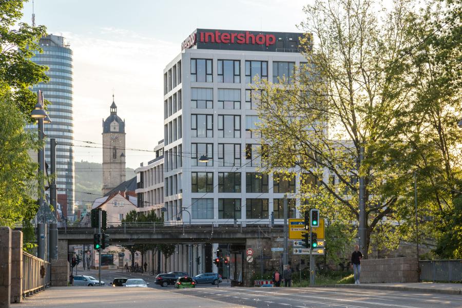 Stadtansicht mit weißen Bürogebäude mit zahlreichen grauen Fenstern, im Hintergrund weitere Häuser und Kirchturm, links und rechts grüne Bäume