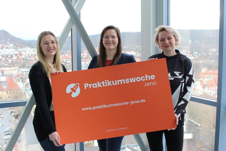 Drei stehen vor großer Fensterfront, stehen nebeneinander und halten oranges Schild mit weißer Schrift