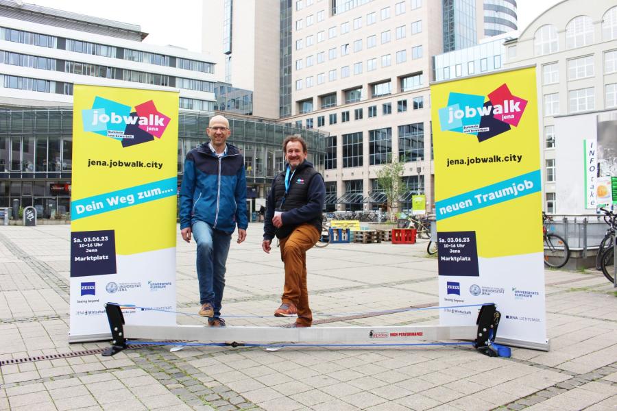 zwei gelb-grüne Roll-ups, dazwischen stehen 2 Männer in legerer Kleidung, im Vordergrund ist Slackline gespannt