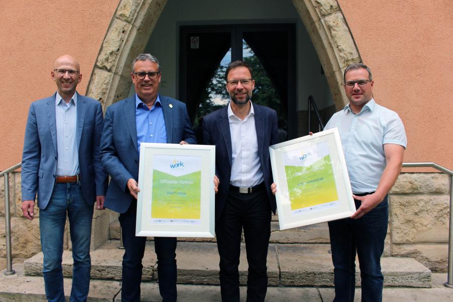 Vier Personen vor einem Eingangstor präsentieren ein Plakat zum i-work Business Award.